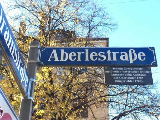 Das Straßenschild der Aberlestraße in München mit einer Zusatztafel, die an den Aufständischen Johann Georg Aberle erinnert, vor einem Baum mit gelben Blättern.